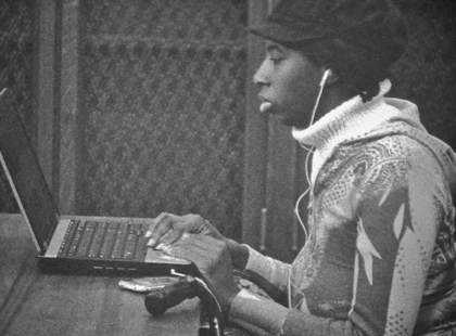 Ute Aurand to be here 2013, film still showing a black woman seated at a laptop and music plugged into her ears.