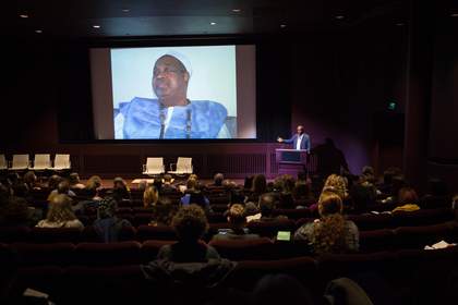 A man stands in front of a projected image onstage in the auditorium at Tate, delivering a lecture to a conference audience