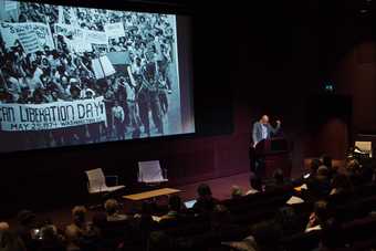 Russell Rickford speaks in front of an audience in the Clore Auditorium at Tate Britain; behind him is a screen showing a photograph of an African Liberation Day protest march in 1974 in Washington, D.C.