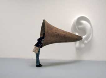 John Baldessari Beethovens Trumpet With Ear Opus 127 2007 a large white ear is mounted on the gallery wall with an ear trumpet attached a visitor has their head inside the trumpet