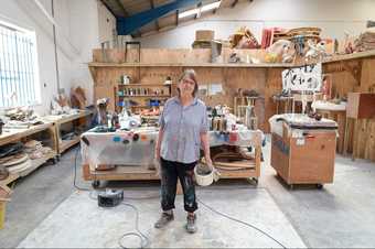 Phyllida Barlow stands in her art studio, with wooden workbenches behind her and wooden shelving on the back wall.
