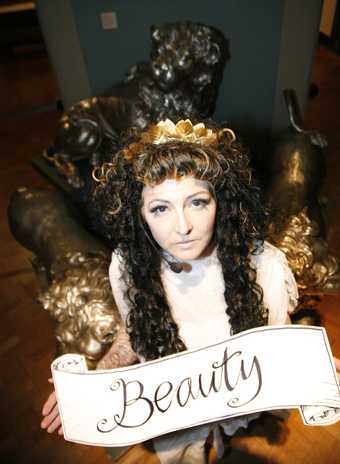 Photograph of a long haired person wearing a crown holding a banner which reads 'Beauty'