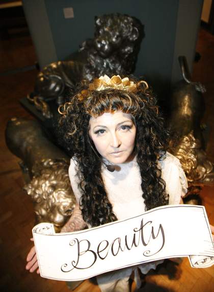 Photograph of a long haired person wearing a crown holding a banner which reads 'Beauty'