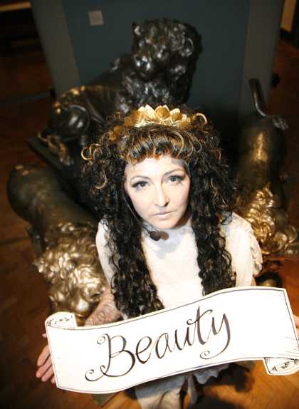 Photograph of a long haired person wearing a crown holding a banner which reads 'Beauty'