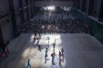 Chain of dancers in bright leotards being watched by a crowd in shadows