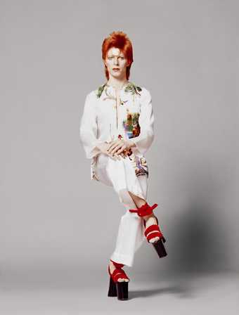 Man with dyed red hair pretends to sit crossed legged whilst standing. He is wearing a white embroidered suit and Japanese style red and black platform shoes.