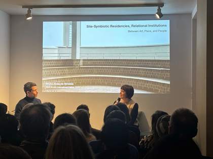 Two people in conversation on a stage with a projected image reading 'Site-Symbolic Residencies, Relational Institutions'