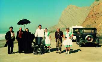 A group of people stand and pose in a sunny mountain landscape including two in nurses uniforms and a man pushing an empty wheelchair