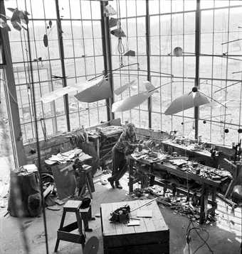 Black and white photograph of Alexander Calder standing in his studio with one of his mobiles hanging above it