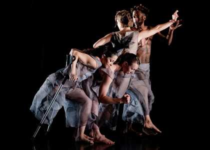 4 dancers are on an all-black stage. They wear loose-fitting sheer grey clothes. The 2 female dancers in front are side-on and crouched down, bending at the knee and upper bodies tilted forward. The front-most dancer holds 2 crutches.