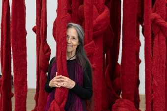 Artist Cecilia Vicuna standing in front of thick strands of red wood hanging from the ceiling.
