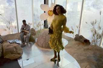 Woman wearing distinctive yellow dress in front of a sculpture of a flower.  Person playing guitar in is sat in the background.