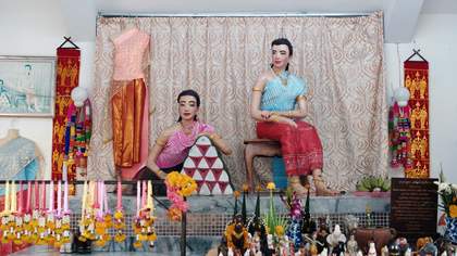 Two human sized dolls sit in a stage set which looks like Bollywood living room