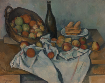 A basket of apples that have tumbled across a cloth on a table, there is also bread and a wine bottle on the table.