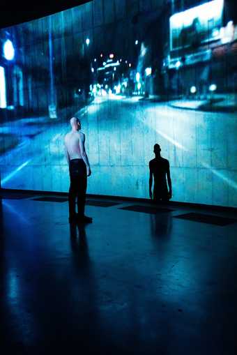 Figure stoo facing the wall in a large open space. A blue-hued projection falls over the figure and onto the wall.