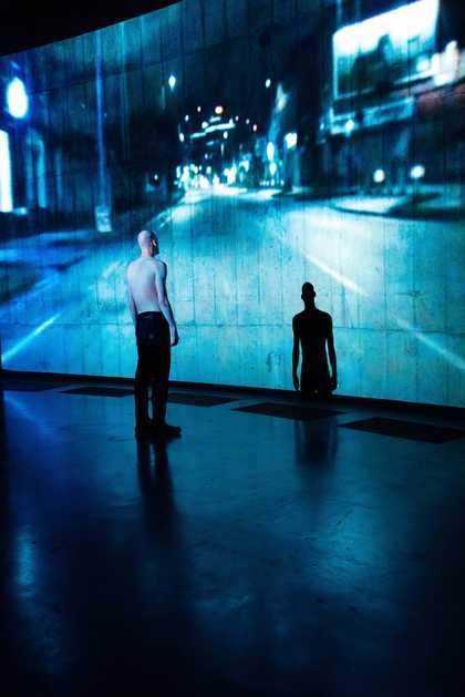 Figure stoo facing the wall in a large open space. A blue-hued projection falls over the figure and onto the wall.