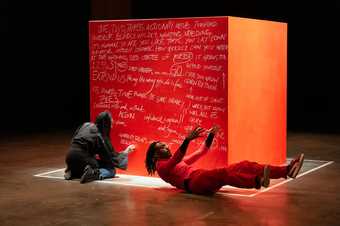 two figures on the floor in front of a large red cube