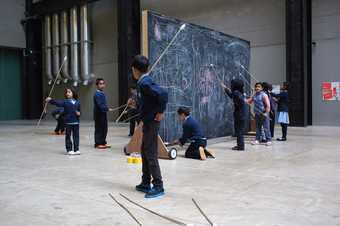 A group of school kids in uniform draw on a giant black board in the turbine hall.