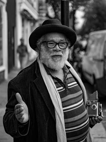 A man looks off beyond us wearing a dark hat and glassess and he has a white beard. He holds an old camera. The photo is black and white.