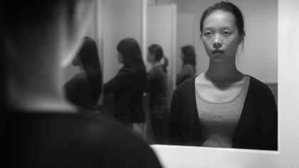 view of a woman looking at her reflection in a mirror. her hair is pulled by and she is dressed plainly.