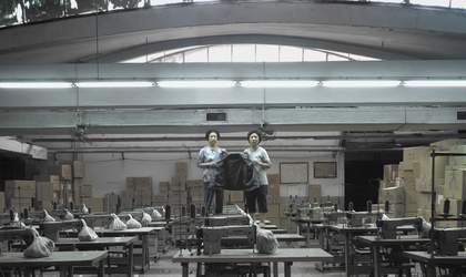 Two mid-age Taiwanese women stand on top of a table on an abandoned garment factory, surrounded by sewing machines and boxes. They hold an empty jacket, gloomily looking to the camera.