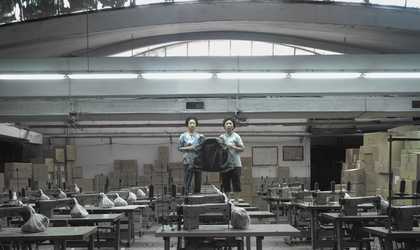 Two mid-age Taiwanese women stand on top of a table on an abandoned garment factory, surrounded by sewing machines and boxes. They hold an empty jacket, gloomily looking to the camera.