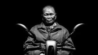 black and white high contrast image of a man sat on a motorbike. only the handles are visible and the background is entirely black.