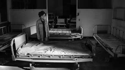 black and white high contrast image of a woman stood in a room of empty beds. it looks like an abandoned asylum or hospital.