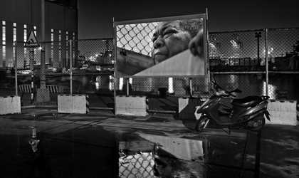 black and white image of a harbour cut off by a metal fence. there's a sign stretched across the fence which shows a portion of a man's face