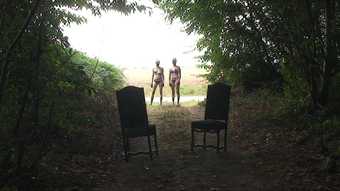 Colour photograph of two chairs in a forest with two figures standing in the distance