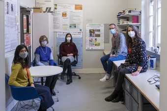 Five people pose in an office, some sitting on chairs, some perched on the edge of a desk, all looking towards the camera. They are distanced and wearing face masks to help prevent the spread of Covid-19.