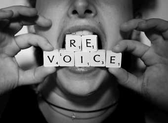 Black and white photograph showing a close up of a person holding Scrabble tiles between their teeth that spell revoice