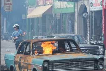 an armed policeman walks behind a car on fire