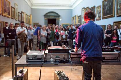 A DJ standing at a DJ booth in front of a crowd of people in one of the galleries at Tate Britain