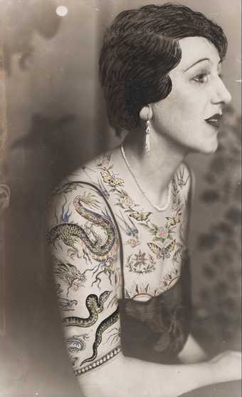 Black and white photograph of a woman sitting in profile, the bust dressed in a blouse made of tattoo patterns drawn on the photograph