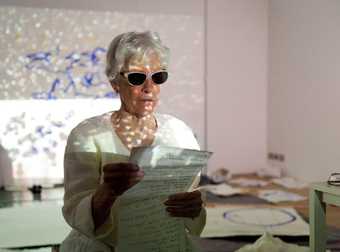 A woman wearing dark glasses reads from paper, from Joan Jonas's Draw Without Looking