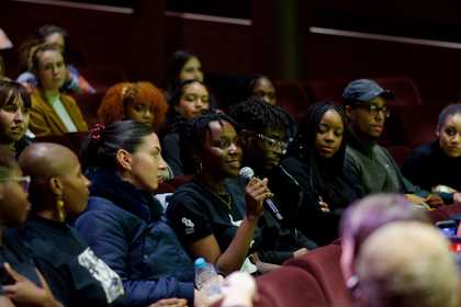 A group of young people in rows of seating in Tate's cinema. The focus is on a person holding a mic in the centre of the of the image