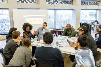 A group of people sit around a large table in a conference room. They are listening to a woman speak. Behind her a large whiteboard reads 'Our data?'.