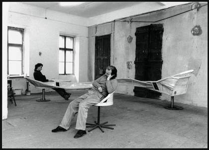 Black and white photograph of two men sat on a piece of furniture featuring three seats joined together