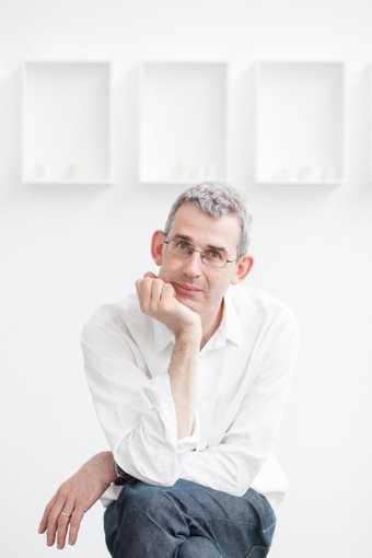 Portrait of Edmund de Waal sitting on a stool in a gallery space with his hand resting on his chin