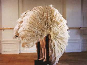 Large featured fan is open entirely covering the figure inside. All the is visible is a pair of legs wearing ballet shoes.