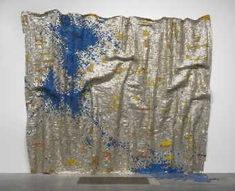 Colour photograph of a square sheet of fabric made of blue, silver and gold coloured squares. The fabric is hanging and its pattern depicts a splash of ink. Made by El Anatsui