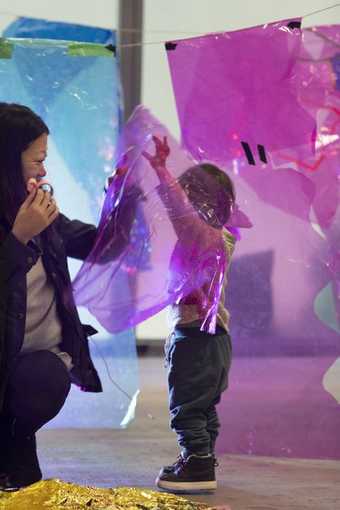 A child and parent play in a making workshop, surrounded by translucent cellophane