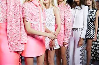 Five blond, white models stand with one coloured model. The outfits are mainly pink, with heavy flowered applique and miniskirts.