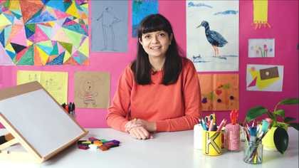 A woman sits facing the camera with an orange jumper on, next to her is a drawing board and behind her are colourful artworks made by children