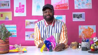 a man sits against a pink backdrop with artworks on paper behind him on the wall, holding a sensory sculpture