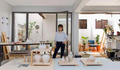 The artist is standing in her studio, facing the camera, with unglazed ceramic objects in front of her.