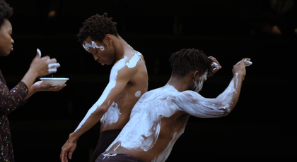 A still image of two dancers covered in white body paint beside a woman with white paint on her hands
