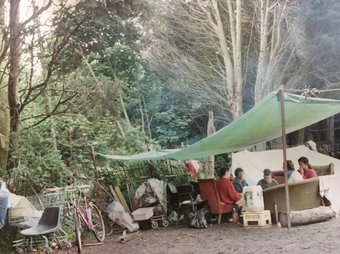 An archival photograph from the 1980s showing a scene of a campsite in woodland. 5 women are sitting on sofas and armchairs under a makeshift awning.
