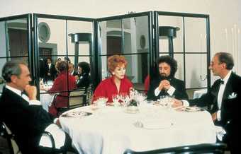 Three men in suits sit at a white clothed table with a red haired woman wearing a red suit sat in between them. They appear to be talking and there are many empty wine glasses on the table. The table is in front of a mirrored screen.